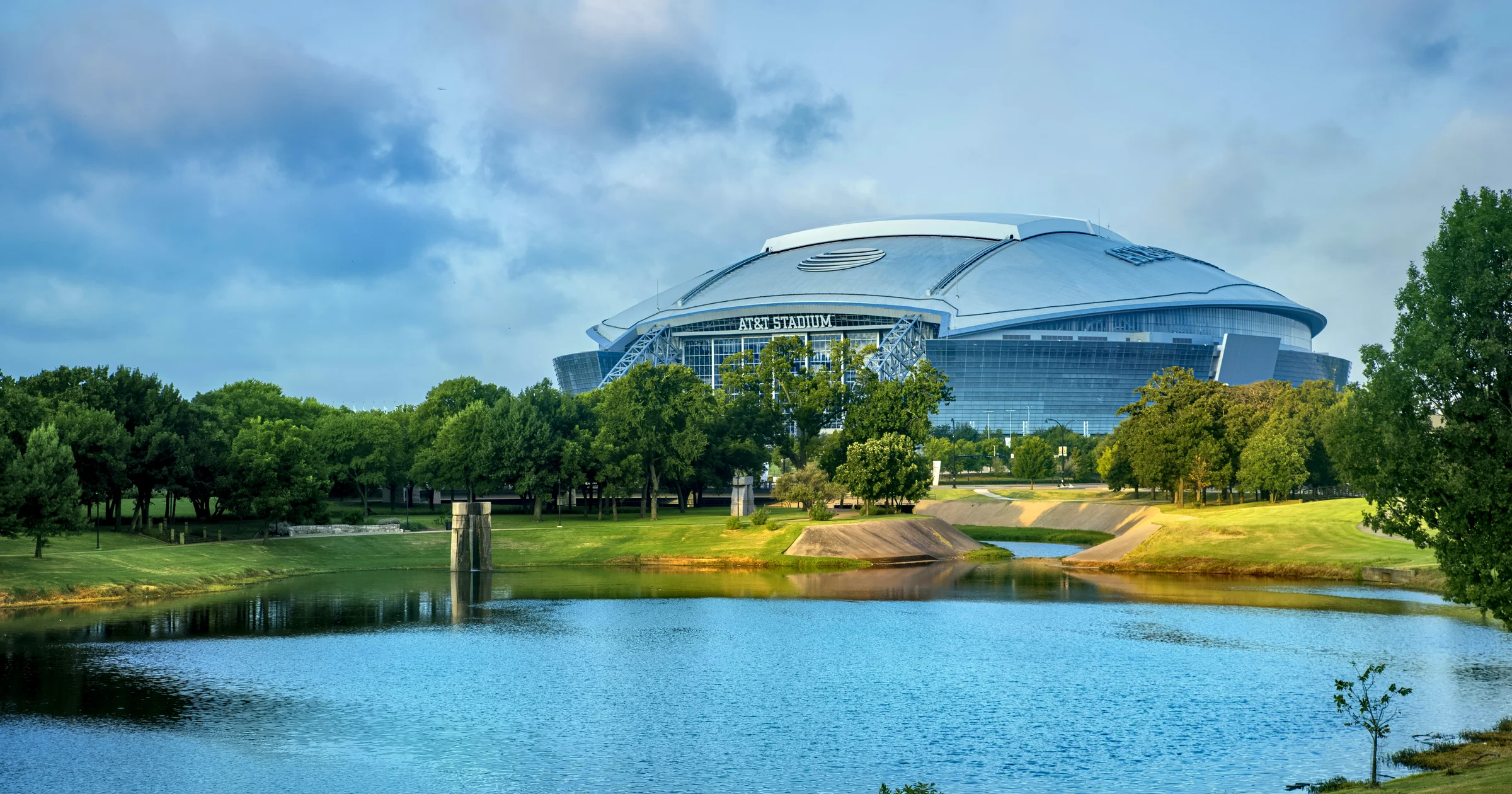 AT&T Stadium in Arlington, Texas rising behind green trees and a calm blue pond under a cloudy summer sky