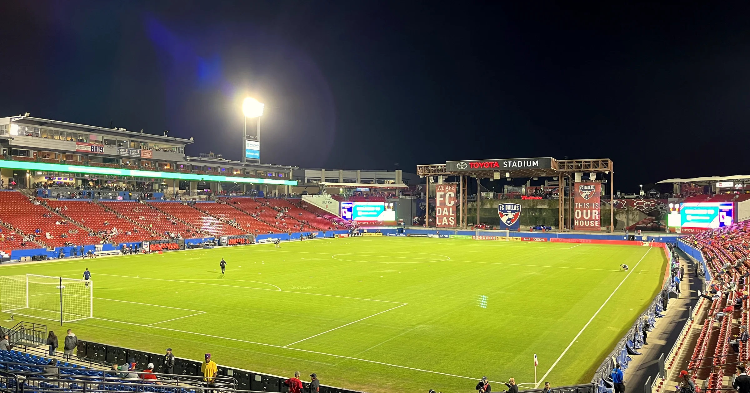 Toyota Stadium in Frisco, Texas at night, floodlights shining over the green pitch and red stands
