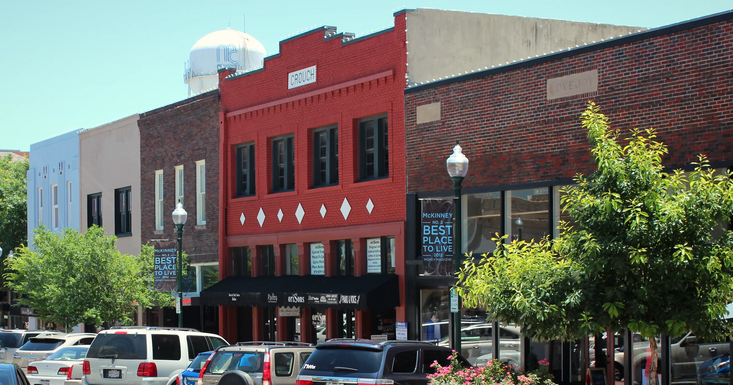 Red and brown brick storefronts with lamp posts and leafy trees on the historic downtown square in McKinney, Texas